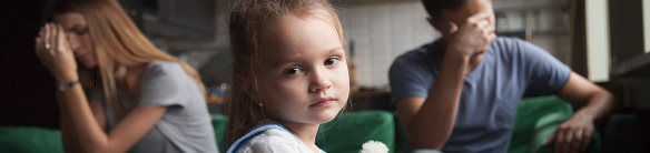 A young child in the centre of the frame looks at the camera. Two parents are folded over in the background. 